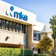 The MKS Instruments building, with its prominent blue logo, stands tall under a clear sky. An American flag waves proudly nearby, symbolizing innovation and progress.