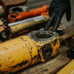 A gloved hand inspects worn hydraulic industrial cylinder mounts.