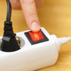 Finger presses a red switch on a white power strip, turning on what is a shared device for multiple plugs.