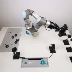 A robotic arm from Adapta Robotics is positioned over a table with various components, ready for assembly or inspection.