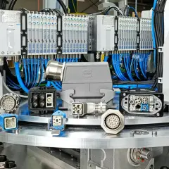 A collection of industrial connectors, including a large grey HARTING connector, are displayed on a metallic surface, with a background of pneumatic valves and wiring.