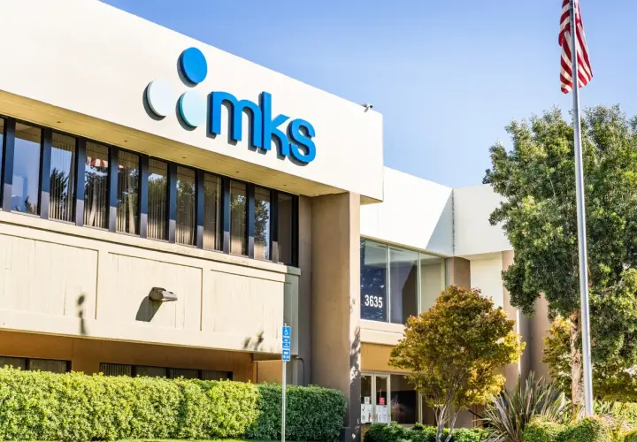 The MKS Instruments building, with its prominent blue logo, stands tall under a clear sky. An American flag waves proudly nearby, symbolizing innovation and progress.
