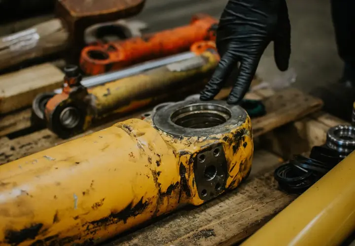 A gloved hand inspects worn hydraulic industrial cylinder mounts.