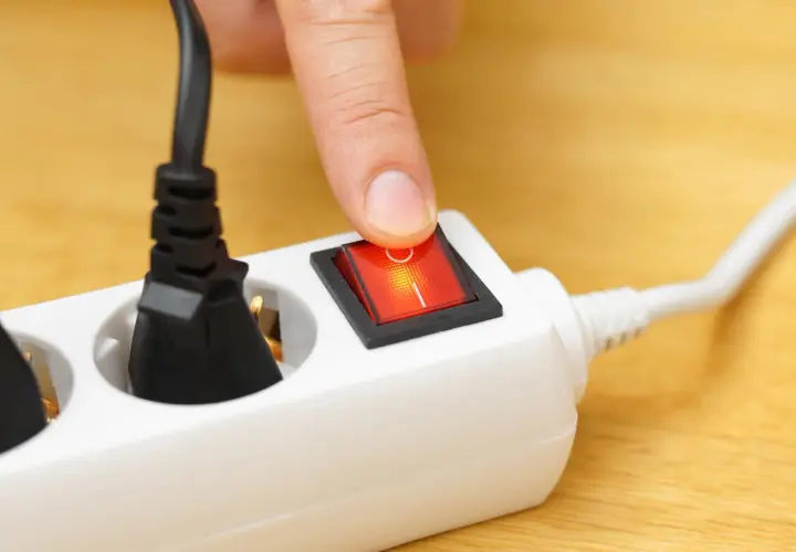 Finger presses a red switch on a white power strip, turning on what is a shared device for multiple plugs.
