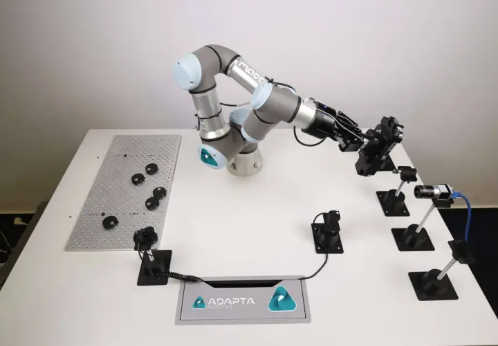 A robotic arm from Adapta Robotics is positioned over a table with various components, ready for assembly or inspection.