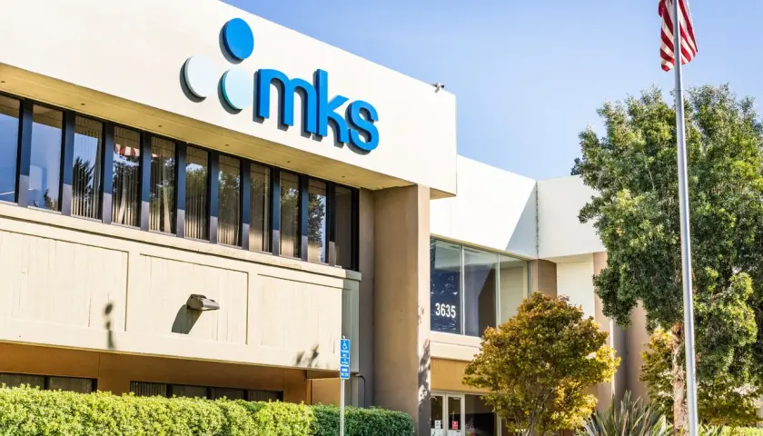The MKS Instruments building, with its prominent blue logo, stands tall under a clear sky. An American flag waves proudly nearby, symbolizing innovation and progress.