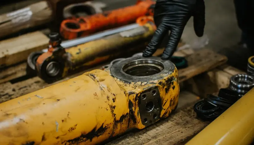 A gloved hand inspects worn hydraulic industrial cylinder mounts.