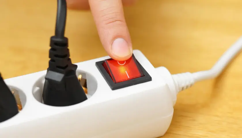 Finger presses a red switch on a white power strip, turning on what is a shared device for multiple plugs.