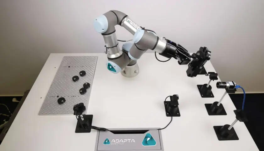 A robotic arm from Adapta Robotics is positioned over a table with various components, ready for assembly or inspection.