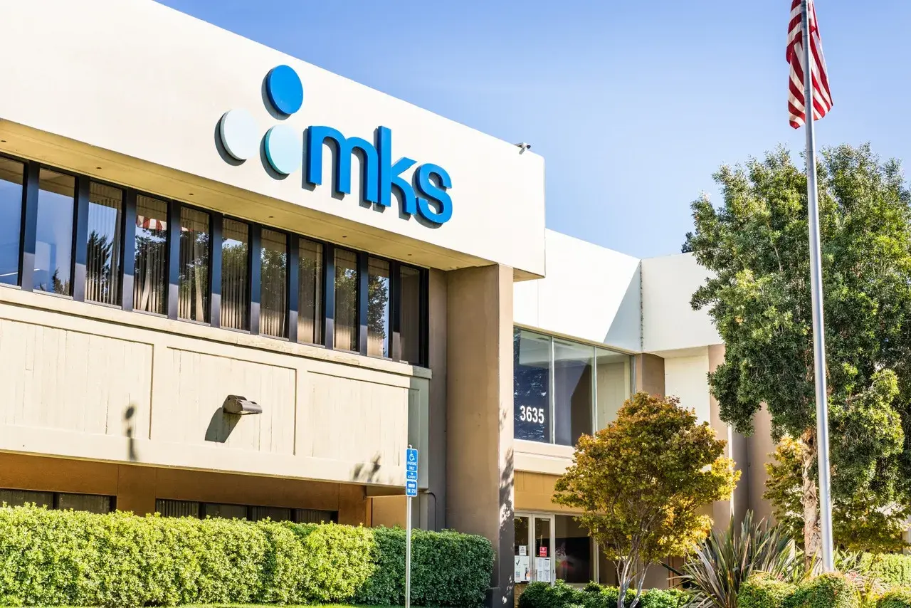 The MKS Instruments building, with its prominent blue logo, stands tall under a clear sky. An American flag waves proudly nearby, symbolizing innovation and progress.