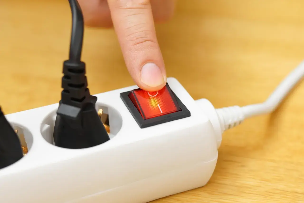 Finger presses a red switch on a white power strip, turning on what is a shared device for multiple plugs.