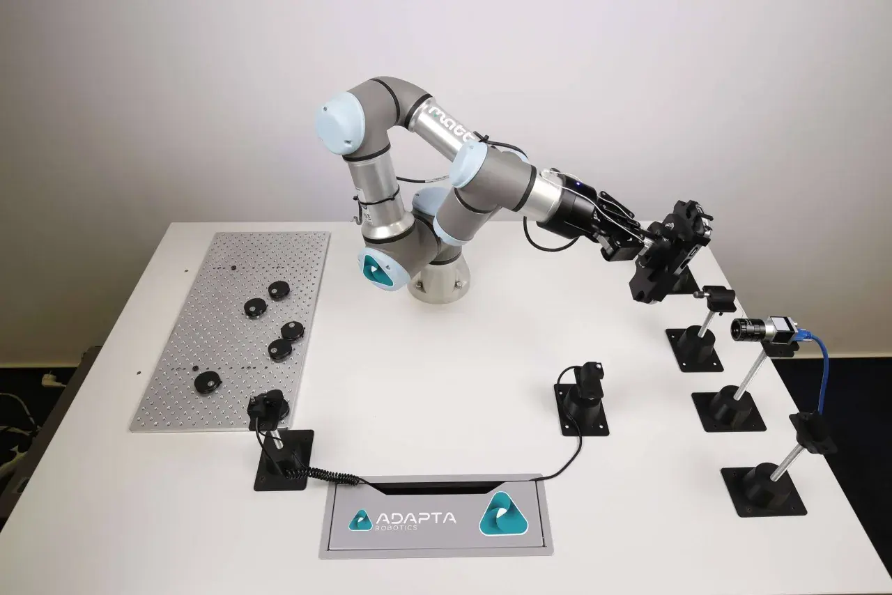A robotic arm from Adapta Robotics is positioned over a table with various components, ready for assembly or inspection.