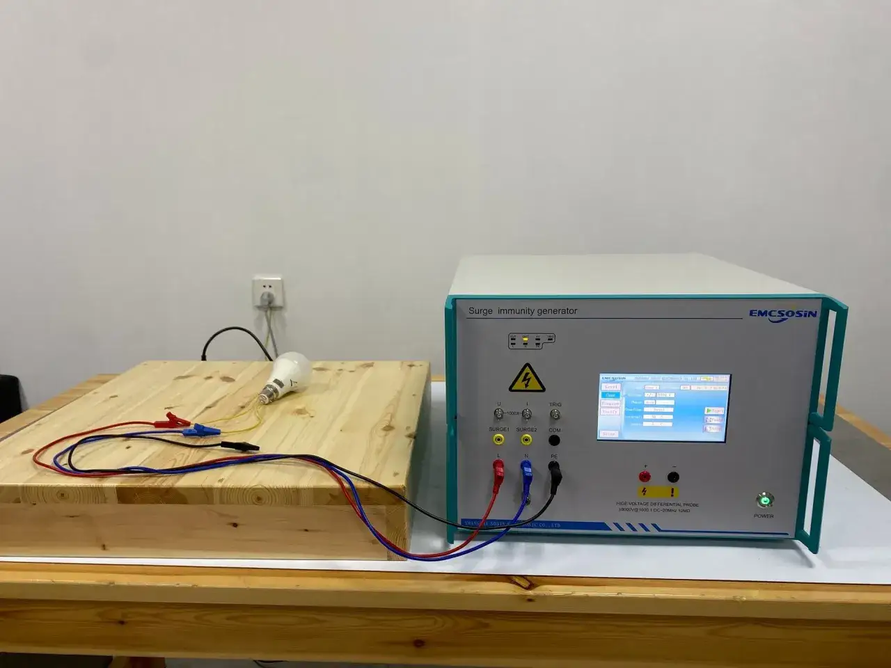 A surge immunity generator is set up for surge testing. A lightbulb on a wooden block is connected to the device, ready to withstand electrical surges.