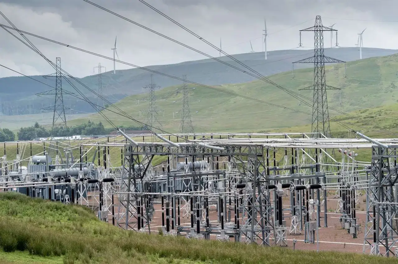 A substation with complex electrical equipment, power lines, and wind turbines on a hilly landscape. This scene illustrates the infrastructure needed to transmit electricity, often measured in kilovolts.
